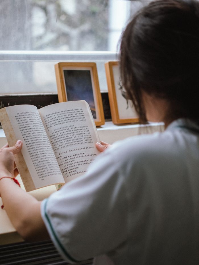 A woman sits indoors reading a book with Chinese text near a window, creating a peaceful ambiance.